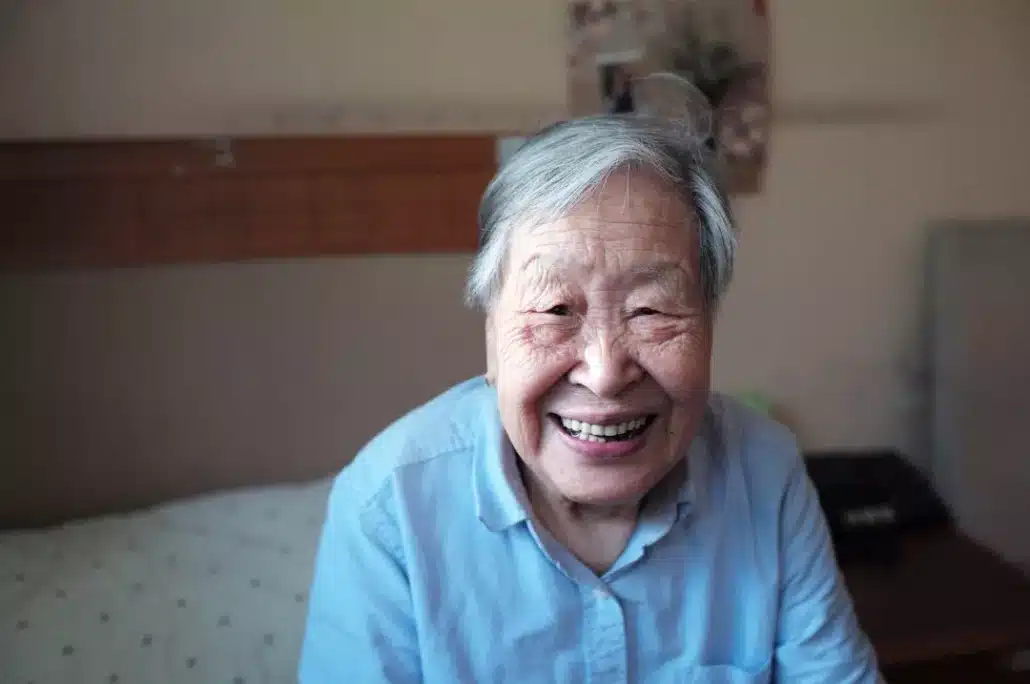 Old woman in blue shirt sitting on a bed in a residential care facility.