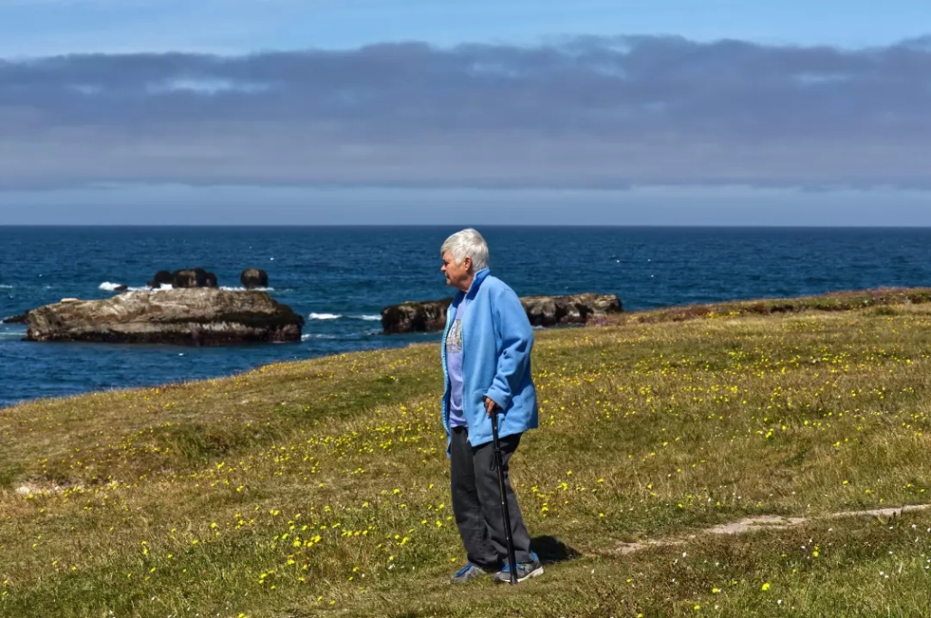Old woman in blue jacket standing in the grass near the ocean.