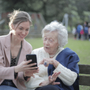 Mother and daughter sitting on a wooden bench in the park while using a phone.