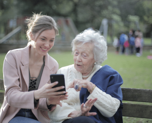 Mother and daughter sitting on a wooden bench in the park while using a phone.