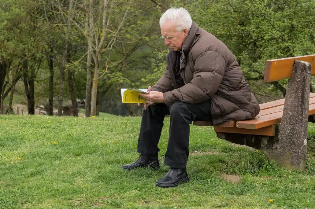 Old man reading a book while sitting on a wooden bench.