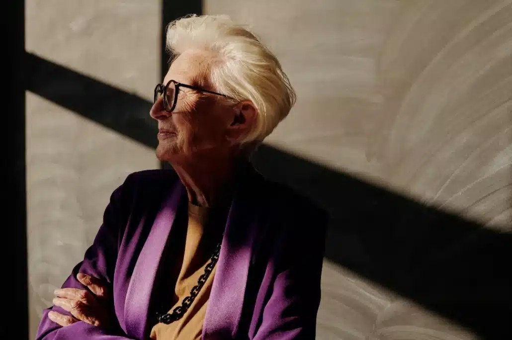 Old woman in purple blazer wearing eyeglasses.