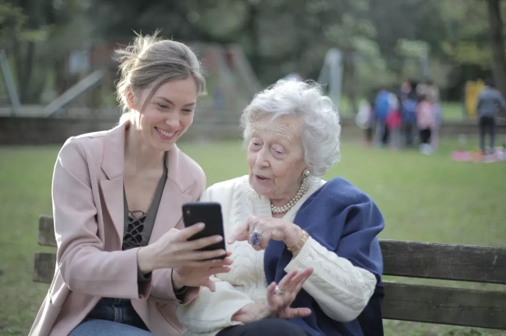 Woman teaching an old woman how to use a cellphone while sitting on a wooden bench.