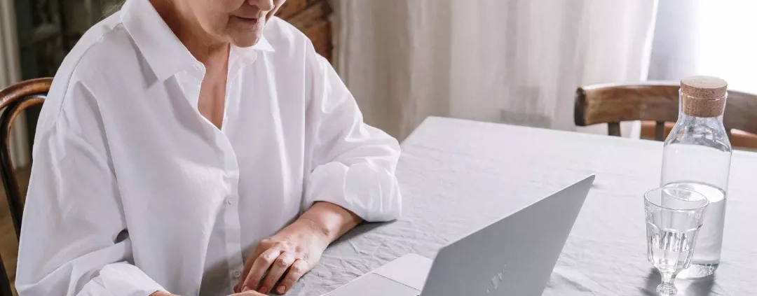 An elderly woman in white long sleeve using a laptop.