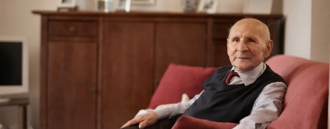 Old man in formal suite sitting on a red sofa.