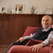 Old man in formal suite sitting on a red sofa.